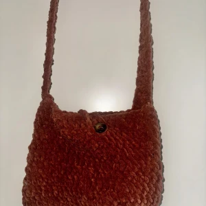 Handvirkad axelväska med knapp - Handvirkad axelväska i dubbelstolpar med fluffig känsla. Väskan har en enkel design med en bred axelrem och stängs med en stor fastsydd knapp framtill. Perfekt storlek för mobil, plånbok och småprylar. Två extra knappar skickas med!