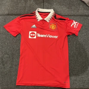 Manchester United Adidas matchtröja M - Köpt på stadium, nypris 1000kr. Röd Manchester United fotbollströja från Adidas med svart-vita detaljer och klubbmärke på bröstet. Klassisk vit krage med röd-svart mönster, svarta axelränder och TeamViewer-tryck. Tillverkad i lätt och ventilerande material, perfekt för match eller träning.