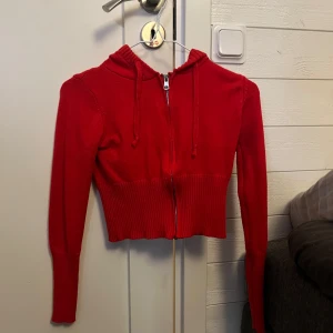 Röd croppad hoodie med dragkedja - Snygg röd croppad hoodie med dragkedja framtill och huva med snörning. Tröjan har långa ärmar och bred ribbad nederkant som ger en cool passform. Perfekt för dig som gillar en trendig och bekväm stil.