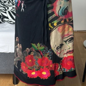 Desigual midi kjol - Unik svart midi kjol från Desigual x Cirque du Soleil med färgstarka tryck av ansikten, rödblommigt mönster och zebraränder. Kjolen har snörning i midjan och coola detaljer som broderade blommor och konstnärliga motiv. Perfekt för dig som gillar statement-plagg.