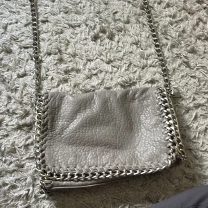 Grå axelväska med kedjedetaljer från Tiamo - Snygg grå axelväska från Tiamo i skinnimitation med grov kedja som axelrem och dekorativa kedjedetaljer runt kanterna. Väskan har dragkedja både på baksidan och inuti, samt ett rymligt huvudfack. Perfekt för dig som gillar en cool och trendig look.