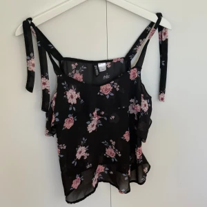 Blommig svart linnetopp från H&M Divided - Svart linnetopp från H&M Divided med rosa och ljusblått blommönster. Toppen har smala axelband med knyt och volangdetaljer längs sidorna. Tunt och luftigt material, perfekt för varma dagar. Snygg och feminin stil som passar till jeans eller kjol.
