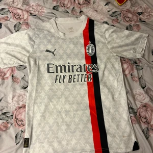 AC Milan vit bortatröja Puma - AC Milan bortatröja från Puma i vitt med subtilt geometriskt mönster, röd och svart rand längs framsidan och broderad klubbmärke. Tryckta sponsorer på bröst och rygg. Tillverkad i lätt och ventilerande polyester med korta ärmar.