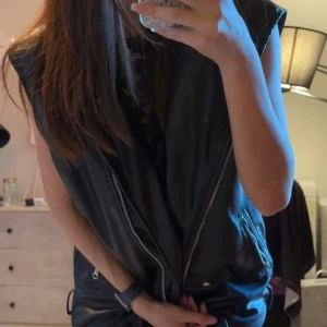 Svart oversized skinnväst med dragkedjor - Säljer en svart oversized väst i skinnimitation med coola dragkedjor på sidorna och fickor med knappdetaljer. Västen har breda axlar och en loose fit som ger en edgy vibe. Perfekt att slänga över en hoodie eller t-shirt för en streetstyle-look. Storlek S