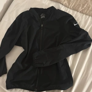 Svart Nike vindjacka Dri-Fit - Svart vindjacka från Nike. Jackan har hel dragkedja framtill, lång ärm och en stilren look med Nike-logga på ena ärmen. Perfekt för aktiva dagar eller när du vill ha en sportig vibe.