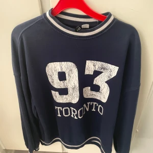 Mörkblå sweatshirt med tryck '93 Toronto' - Mörkblå sweatshirt från Gina Tricot med stort vitt tryck '93 Toronto' på bröstet. Tröjan har rund hals med vit och blå rand, samt matchande rand vid nederkanten. Långärmad och i mjuk bomullsblandning, perfekt för en chill och sportig look.