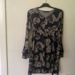 Blommig svart klänning  - Supersnygg svart kortklänning från Soul River med lila och grått blommönster. Klänningen har v-ringning och långa trumpetärmar. Perfekt för dig som gillar en feminin och trendig stil.