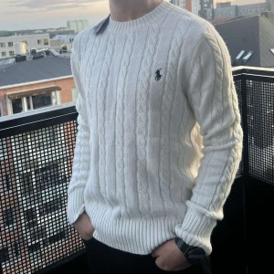 Polo ralph lauren sweater - Kabelstickad vit tröja från Polo Ralph Lauren med klassisk rund hals och broderad marinblå logga på bröstet. Tröjan är långärmad och har ribbade muddar och nederkant. Perfekt för dig som gillar stilren och tidlös design.