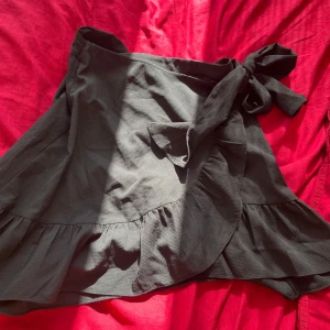 Svart omlottkjol med volang - Supersnygg svart kort omlottkjol med volangkant och knyt i midjan. Säljer då den inte kommer till användning 