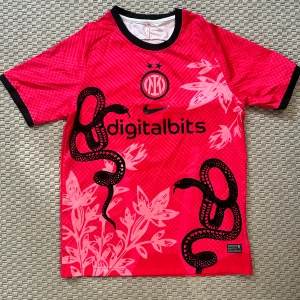 Inter Milan x Nike rosa fotbollströja - Inter Milan x Nike matchtröja i stark rosa med svarta detaljer och coolt ormmotiv samt blommor på framsidan. Kortärmad modell med svart krage och ärmslut. Tillverkad i lätt och ventilerande funktionsmaterial, perfekt för fotbollsplanen.