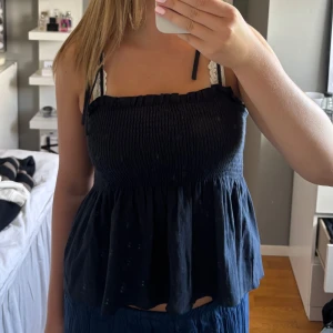 Mörkblå smockad linnetopp med knytband - Supersöt mörkblå linnetopp med smockad överdel och volangig nederdel. Tunna axelband som knyts på axlarna för en personlig touch. Perfekt till varma dagar och enkel att styla med jeans eller kjol.