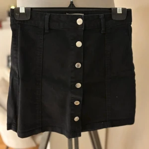 Svart jeanskjol med knappar, Gina Tricot - Svart kort jeanskjol från Gina Tricot med raka linjer och snygga silverfärgade knappar framtill. Klassisk femficksmodell med hällor för bälte. Perfekt att matcha med en cool t-shirt eller stickad tröja.