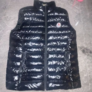 Svart dunväst från Moncler - Snygg svart dunväst från Moncler med glansig finish och klassisk logga på bröstet. Västen har ståkrage, två dragkedjefickor och quiltad design. Perfekt att slänga över en hoodie för en clean look.diskutera priset .