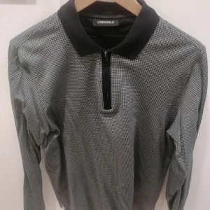 Golf/sports pikétröja Lagerfeld - Snygg långärmad pikétröja från Lagerfeld med svart krage och diskret rutigt mönster i svart och vitt. Tröjan har half zip-dragkedja framtill och klassisk passform. Perfekt för dig som gillar stilrena och tidlösa plagg. Elastisk 100% bomull för sports/golf.