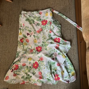 Blommig omlottkjol i satin - Superfin lång omlottkjol i satin med blommigt mönster i rosa, rött, gult och grönt på vit botten. Kjolen har volangkant och knyts i midjan med ett band. Perfekt för sommarens alla tillfällen och ger en romantisk vibe. Resår i midjan.