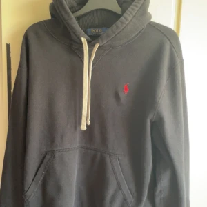 Svart hoodie från  Ralph Lauren - Svart hoodie från Polo Ralph Lauren med klassisk röd logga broderad på bröstet. Tröjan har huva med vita snören och en stor magficka framtill. Perfekt för en chill och stilren look.