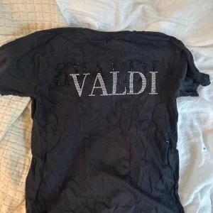 Svart t-shirt från Bvaldi med strass - Svart t-shirt från Bvaldi med korta ärmar och rund hals. Framsidan har en stor VB-logga i glittrande strass och baksidan texten 'BVALDI' i strass. Skön passform och perfekt för dig som gillar statement-plagg.