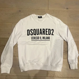 Vit sweatshirt från Dsquared2 - Säljer en vit sweatshirt från Dsquared2 med svart tryck framtill där det står 'DSQUARED2 CERESIO 9, MILANO'. Tröjan har rund halsringning, långa ärmar och ribbade muddar. Perfekt för dig som gillar stilrena och trendiga plagg.