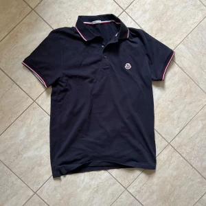 Svart Moncler pikétröja med röd/vit kant - Svart pikétröja från Moncler med klassisk krage och knäppning framtill. Röd och vit randig detalj på krage och ärmslut. Broderad Moncler-logga på bröstet. Perfekt för dig som gillar stilrena och exklusiva plagg.