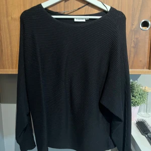 Svart ribbad tröja från Kappahl - En svart långärmad tröja från Kappahl med ribbad struktur. Tröjan har en avslappnad passform och rund halsringning, perfekt för dig som gillar en enkel men stilren look. Passar till allt och är superenkel att matcha med andra plagg.