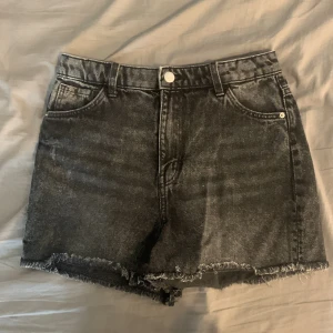 Svarta jeansshorts med fransig kant - Snygga svarta jeansshorts med hög midja och rå, fransig kant nertill. Klassisk femficksmodell med bälteshällor och knappstängning. Perfekta för sommaren och ger en cool, avslappnad vibe.