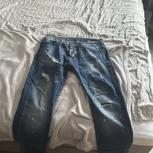 Dsquared2 blå distressed jeans - Snygga blå jeans från Dsquared2 med slitna detaljer, hål och färgstänk för en riktigt edgy look. Klassisk femficksmodell med läderpatch bak i midjan och raka ben. Perfekta för dig som gillar streetstyle och vill sticka ut.