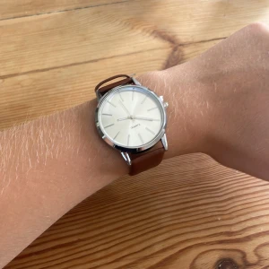 Stilren silverklocka med brunt band  - En klassisk och stilren klocka från Quartz med rund silverfärgad boett och ljus urtavla. Klockan har ett brunt band i syntetmaterial och minimalistisk design utan siffror, perfekt för dig som gillar enkel och clean stil.