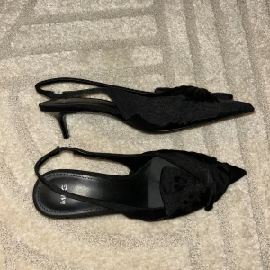 Svarta med rosett - Snygga svarta MNG med elegant rosettdetalj framtill och spetsig tå. Skorna har låg klack och öppen häl med smal rem. Perfekta för dig som vill ha en stilren och trendig look.