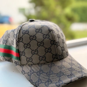 Fet Gucci keps - Snygg keps från Gucci med klassiskt GG-monogram i mörkgrått på beige botten. Kepsen har en grön och röd rand på sidan och justerbar rem baktill. Perfekt accessoar för att lyfta din outfit med lyxig streetstyle.