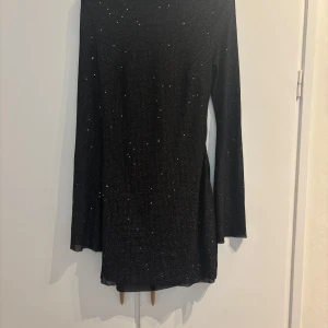 Svart glittrig kortklänning med öppen rygg - Supersnygg svart kortklänning med glittriga detaljer och lång ärm. Klänningen har en djup öppen rygg med snygg drapering och knytband i nacken. Perfekt för dig som vill sticka ut med en elegant och trendig look. Använd endast en gång. Kommer ej ihåg märke