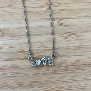 Silverfärgat halsband med LOVE-text - Ett snyggt halsband i silverfärg med texten LOVE där bokstäverna är täckta av glittrande stenar och O:et är format som ett hjärta. Perfekt accessoar för att lyfta din outfit med lite extra bling. Den är från uropenn och har tappat färgen lite så den säljs billigt!!