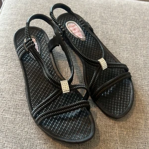 Svarta sandaler med flätade remmar - Svarta sandaler med flätade syntetremmar och dekorativa silvriga detaljer. Platt sula med mönstrad yta för extra grepp. Perfekta för varma dagar och enkel att matcha med olika outfits.