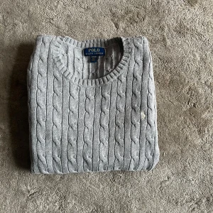 Grå kabelstickad tröja Polo Ralph Lauren - Kabelstickad grå tröja från Polo Ralph Lauren med rund halsringning och diskret broderad logga på bröstet. Klassisk modell med ribbade muddar och nederkant, perfekt för lager-på-lager under kyliga dagar.