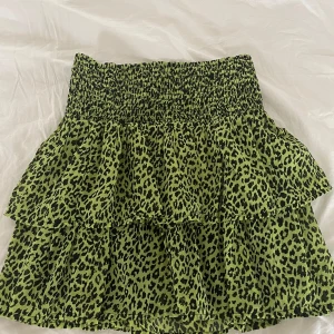 Volangkjol - Supersöt kort kjol i grön med svart leopardmönster och volanger. Kjolen är i storlek 146/153 men jag skulle säga att den passar någon som har runt storlek S. Den har smock i midjan för en bekväm passform och är gjord i ett lätt, luftigt material.