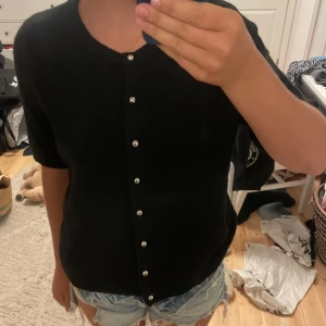Stickad kortärmad kofta - Svart kofta med korta ärmar och rund halsringning. Stängs med snygga pärlknappar framtill. Perfekt att slänga över en topp eller klänning för en chill men ändå stilren look. Det är storlek L men jag har S/M vanligtvis och jag tycker den sitter perfekt på mig!🩷