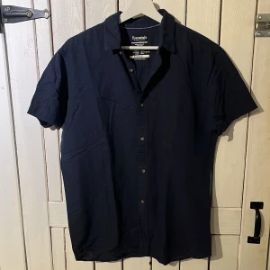 Mörkblå skjorta Jack & Jones Essentials - Snygg mörkblå kortärmad skjorta från Jack & Jones Essentials. Skjortan har klassisk krage, knäppning framtill och är tillverkad i en skön mix av bomull och linne. Perfekt för varma dagar när du vill vara både stilren och bekväm.