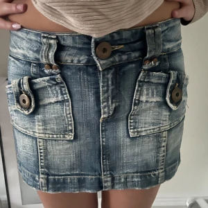 Blå jeanskjol med stora fickor - Säljer en snygg kort jeanskjol i blått med slitna detaljer och två stora fickor framtill med coola knappar. Kjolen har hög midja och markerad söm, perfekt för en trendig och avslappnad look.