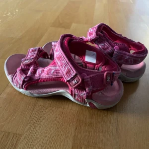 Rosa sandaler med kardborreband - Snygga rosa sandaler med justerbara kardborreband och vit sula. Skorna har ett sportigt utseende och är perfekta för varma dagar. Mönstret är diskret randigt i olika rosa nyanser och materialet är syntet med textil.