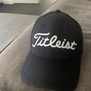 Svart keps från Titleist - Snygg svart keps från Titleist med vit logga framtill. Klassisk böjd skärm och justerbar passform. Insidan har flera Titleist-loggor tryckta längs sömmarna. Perfekt för golf eller en sportig look.