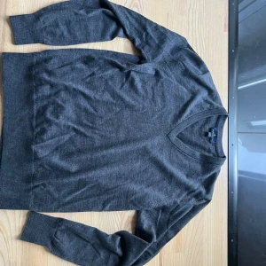 Mörkgrå v-ringad tröja Brooks Brothers - Mörkgrå v-ringad tröja från Brooks Brothers i mjuk och tunn stickad kvalitet. Klassisk modell med långa ärmar och ribbade muddar. Perfekt för lager-på-lager och stilrena outfits. Har ett litet hål i nacken men inget som direkt syns