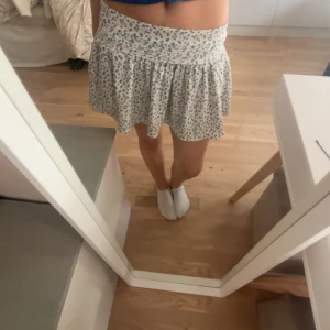 Vit blommig kortkjol med volang - Supersöt vit kortkjol med blått blommönster och bred smock i midjan. Kjolen har volangig nederdel som ger en flowy och luftig känsla.