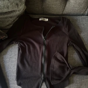 Svart ribbad kofta från Nova - Svart ribbad kofta från Nova med lång ärm och dragkedja framtill. Tröjan har en tight passform och är tillverkad i ett stretchigt material som sitter snyggt på kroppen. Perfekt att slänga över en topp för en chill look.