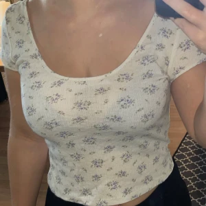 Blommig croppad topp med kort ärm - Supersöt croppad topp i ljusbeige med små lila blommor över hela. Djup rundad urringning både fram och bak, korta ärmar och figurnära passform. Perfekt för vår och sommar, och lätt att matcha med jeans eller kjol.