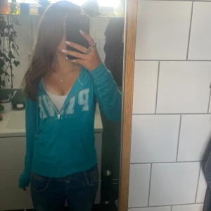 Turkos hoodie från PINK Victoria's Secret - Säljer en turkos hoodie från PINK Victoria's Secret med stor vit logga framtill. Tröjan har dragkedja, huva med snörning och känguruficka. Perfekt för chill dagar och snygg till jeans. Mjuk bomullskänsla och långärmad modell.