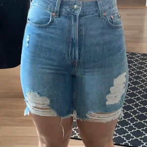 Högmidjade blå jeansshorts med slitningar - Säljer ett par snygga högmidjade jeansshorts från H&M i klassisk blå denim. Shortsen har råa kanter och slitna detaljer framtill för en cool, avslappnad look. Femficksmodell med bälteshällor och dragkedja med knapp. Perfekta för sommaren!