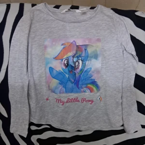 Grå My Little Pony långärmad tröja - Supergullig grå långärmad tröja med stort färgglatt My Little Pony-tryck framtill. Motiv med Rainbow Dash och Twilight Sparkle, plus texten 'My Little Pony' i rosa. Bilden ändras beroende på vilket håll man kollar ifrån. Detta är en barn tröja, för åldern 8-10 år