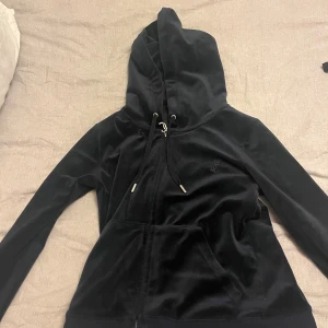 Juicy Couture - Mörkblå hoodie från Juicy Couture i mjuk velour med dragkedja och snörning i huvan. Klassisk logga broderad på bröstet och känguruficka framtill. Perfekt för en chill och trendig look. 