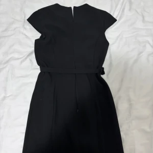 Svart klassisk klänning med bälte - En svart, stilren klänning med bälte i midjan och korta ärmar. Klänningen har en rak, elegant passform och dragkedja bak. Perfekt för dig som gillar en tidlös look med minimalistiska detaljer.