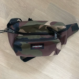 Camouflage magväska från Eastpak - Säljer en snygg magväska från Eastpak i klassiskt camouflage-mönster. Väskan har två dragkedjefack och justerbart band, perfekt för att bära det viktigaste. Materialet är slitstark polyester och färgerna går i grönt, brunt och beige.