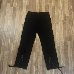 Svarta cargobyxor - Svarta cargobyxor med flera stora fickor på sidorna och baktill. Byxorna har raka ben med dragkedjor och snörning vid bensluten för en cool streetstil. 